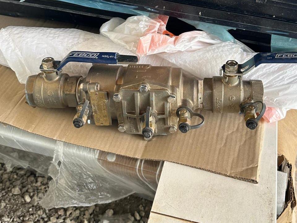 A brass backflow preventer assembly with blue handled shutoff valves and test cocks, resting on cardboard