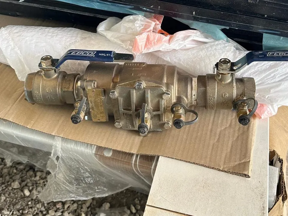 A brass backflow preventer assembly with blue handled shutoff valves and test cocks, resting on cardboard