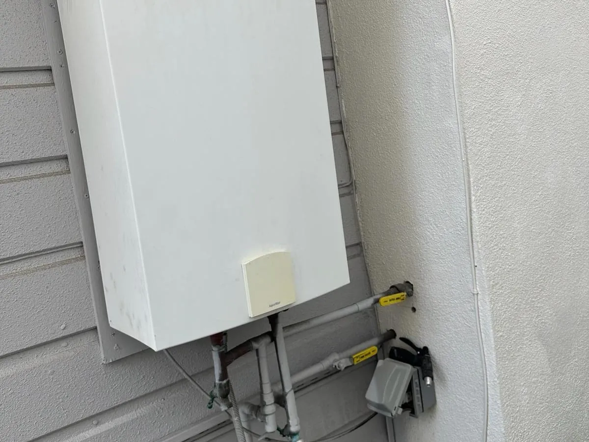 The lower part of a wall mounted outdoor tankless water heater on gray siding, with water lines and yellow handled gas valves connected below it, next to a stucco column
