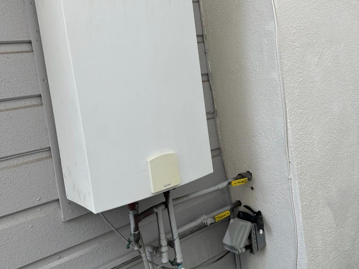 The lower part of a wall mounted outdoor tankless water heater on gray siding, with water lines and yellow handled gas valves connected below it, next to a stucco column