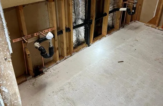 A lower wall opened to the studs showing two black sink drain traps, copper supply stubs on copper brackets and a black drain line running to the stack, above a bare floor