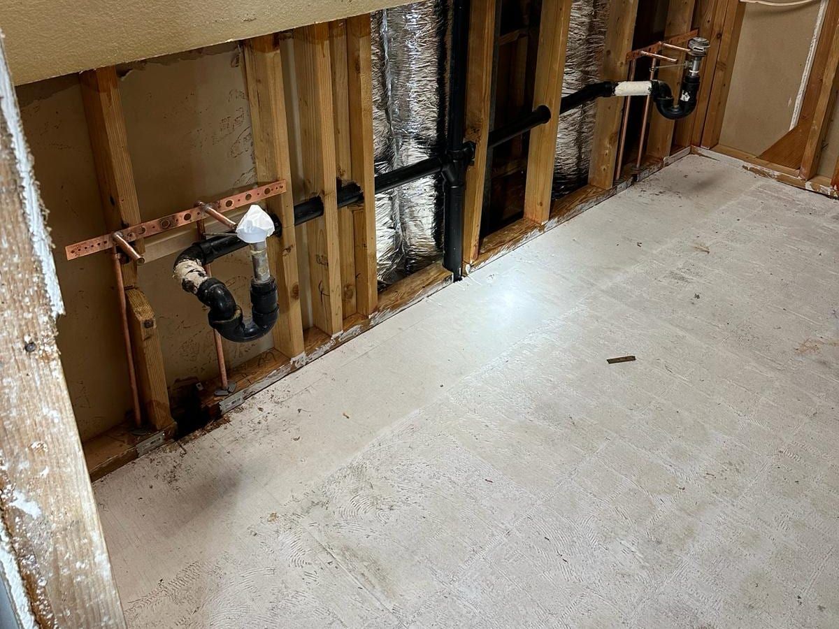 A lower wall opened to the studs showing two black sink drain traps, copper supply stubs on copper brackets and a black drain line running to the stack, above a bare floor