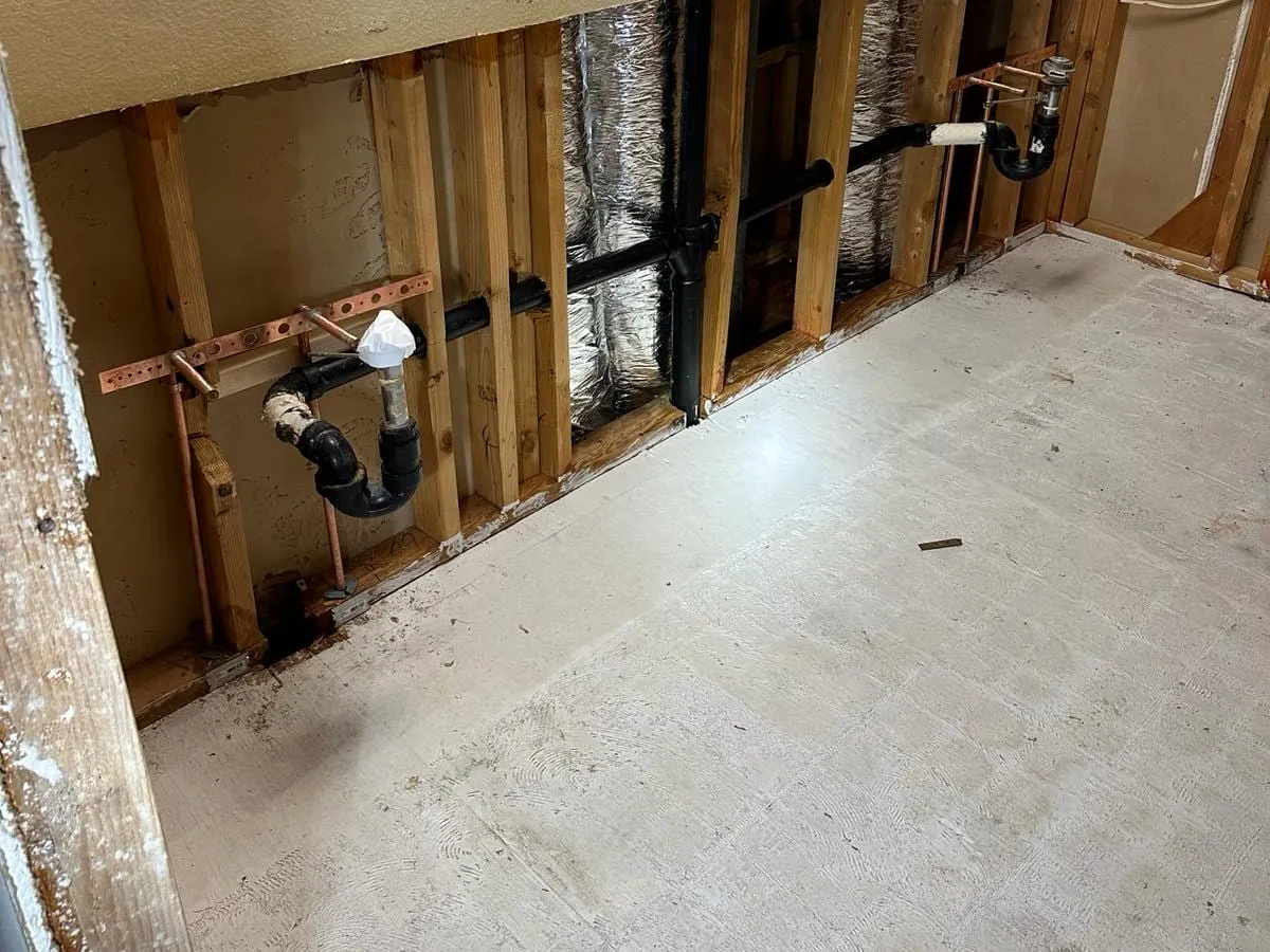 A lower wall opened to the studs showing two black sink drain traps, copper supply stubs on copper brackets and a black drain line running to the stack, above a bare floor