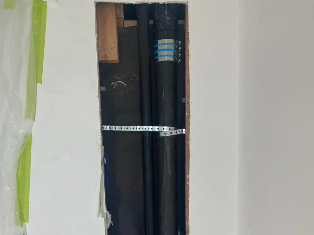 A narrow opening cut in a white wall, showing two black drain stacks joined with banded couplings and held by a metal strap, with plastic sheeting taped beside it