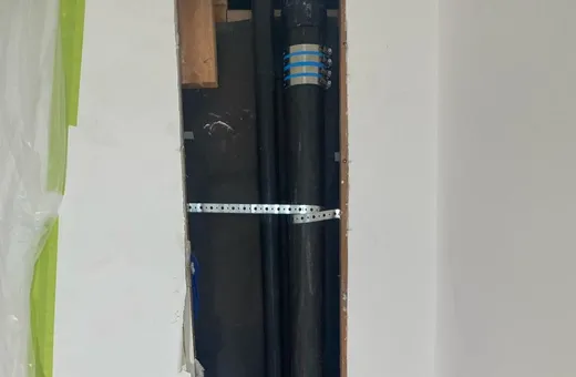 A narrow opening cut in a white wall, showing two black drain stacks joined with banded couplings and held by a metal strap, with plastic sheeting taped beside it