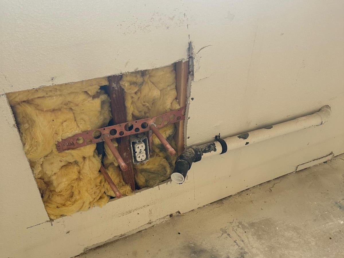 A square opening cut in a drywall wall, showing insulation, copper pipe stubs on a metal bracket and an outlet, beside a white drain pipe with a black coupling running along the wall