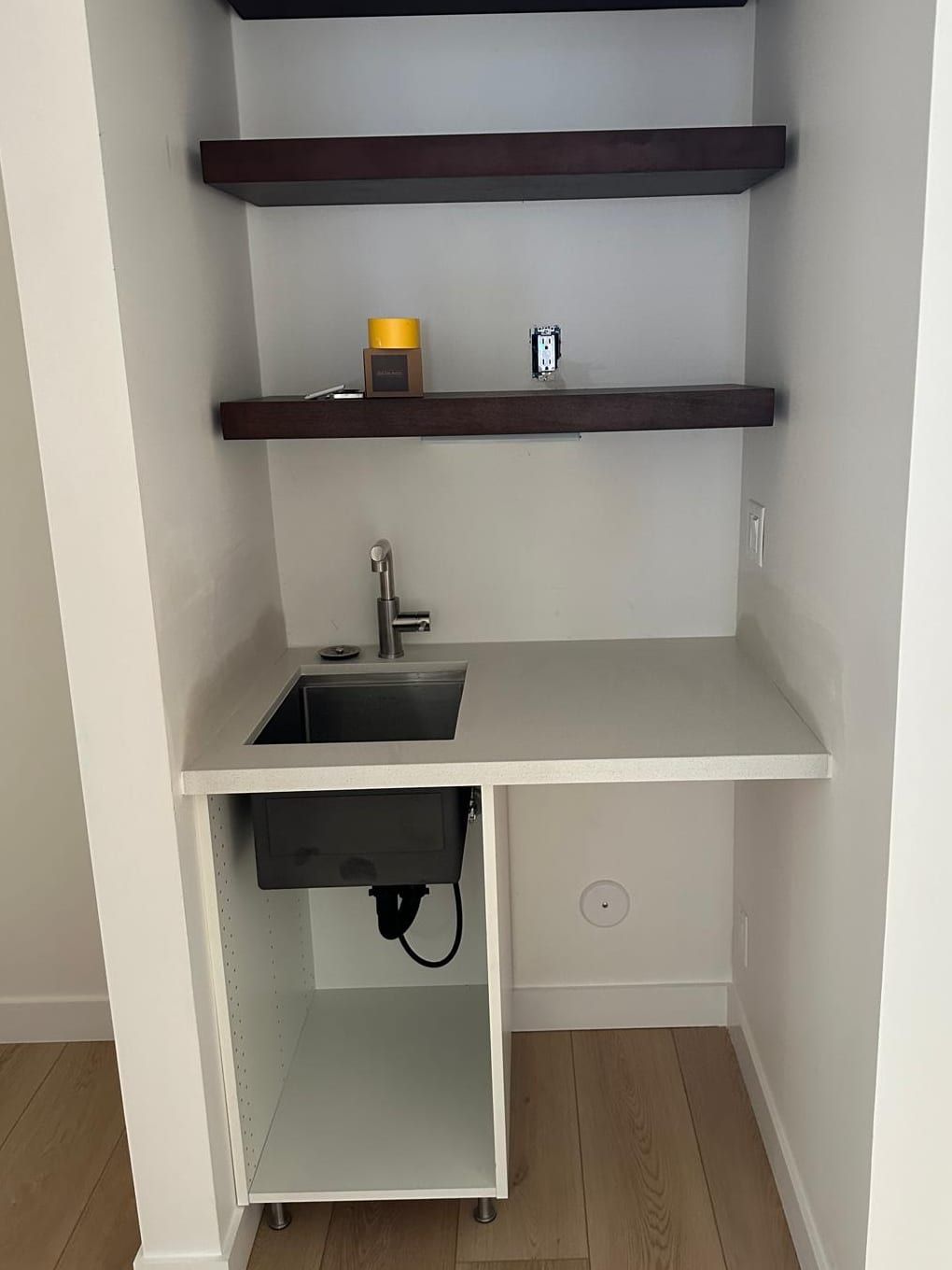 A small wet bar alcove with a stainless undermount sink and a brushed nickel faucet in a white quartz counter, an open white base cabinet below it and dark wood floating shelves above